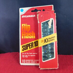 May include: A red and white box of Sylvania Flip Flash Super 10 flashbulbs. The box has a blue dot and says "Qualité Blue Dot: Quality". The box says "S-10 CD" and "10 flashes éclairs". The box also says "For all Flip Flash cameras" and "Pour tous les appareils Flip Flash".