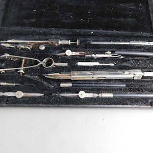 May include: A vintage set of drafting tools in a black leather case. The tools include a compass, a protractor, a ruler, and a pencil.