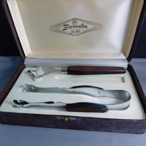 May include: A brown leatherette box with a gold latch, containing a set of three silver bar tools. The tools include a bottle opener, a pair of tongs, and a small ice pick. The box is labelled "Barmates Styled by Glo-Mitt Canada."