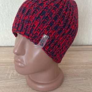 May include: A chunky knit beanie featuring a two-tone pattern of vibrant red and dark charcoal gray yarn. The hat has a ribbed texture and includes a small, rectangular, translucent plastic tag attached with a silver metal rivet on the side.