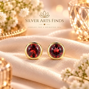 Peut inclure: Une paire de boucles d'oreilles à clous rondes avec des pierres précieuses facettées rouge foncé serties dans des montures en métal doré poli. Le texte SILVER ARTS FINDS JEWELRY FOR A BRIGHTER YOU apparaît au-dessus avec un petit cœur.