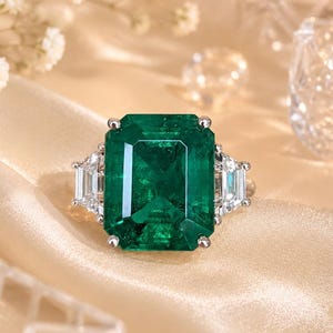 Peut inclure: Une bague ornée d'une grande pierre précieuse verte de taille émeraude rectangulaire, montée sur un anneau en métal argenté. La pierre centrale est flanquée de chaque côté de deux petites pierres claires de taille baguette.
