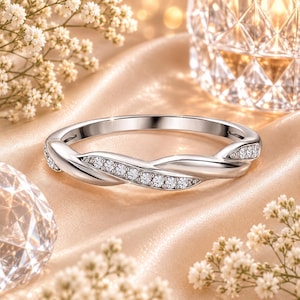 Op de afbeelding: Een zilverkleurige metalen ring met een gedraaid bandontwerp. De ene helft van de band is glad en gepolijst, terwijl de andere helft is ingelegd met een rij kleine, heldere, rondgeslepen stenen die het licht vangen op het gebogen oppervlak.