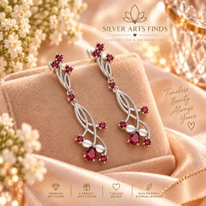 Peut inclure: Une paire de boucles d'oreilles pendantes en argent avec un motif de branche orné de plusieurs petites pierres rouges rondes et d'une pierre rouge en forme de poire à la base. Le texte SILVER ARTS FINDS et Timeless Beauty Always Yours est visible.