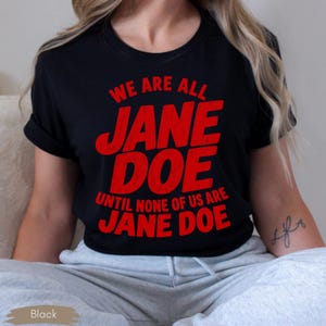 May include: A black short-sleeved t-shirt featuring bold red text that reads WE ARE ALL JANE DOE UNTIL NONE OF US ARE JANE DOE. The lettering has a slightly distressed, textured appearance and is centered on the front of the garment.