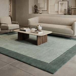 Puede incluir: Un conjunto de sala de estar con un sofá largo de color beige, dos sillones a juego con marcos de metal fino y una mesa de centro rectangular con tapa clara y base de madera oscura. Una alfombra grande con borde verde salvia cubre el suelo.