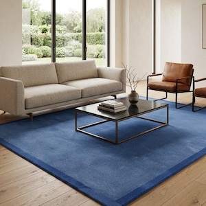 Puede incluir: Una alfombra rectangular grande de color azul profundo sólido con un borde a juego. La alfombra está colocada sobre un suelo de madera clara en una habitación que cuenta con un sofá beige, dos sillones de cuero marrón y una mesa de centro.