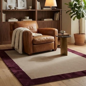 Puede incluir: Una alfombra rectangular que presenta un gran panel central de color beige enmarcado por un borde ancho de color burdeos intenso. La alfombra está colocada sobre un suelo de madera clara, proporcionando una base neutra para los muebles y objetos.