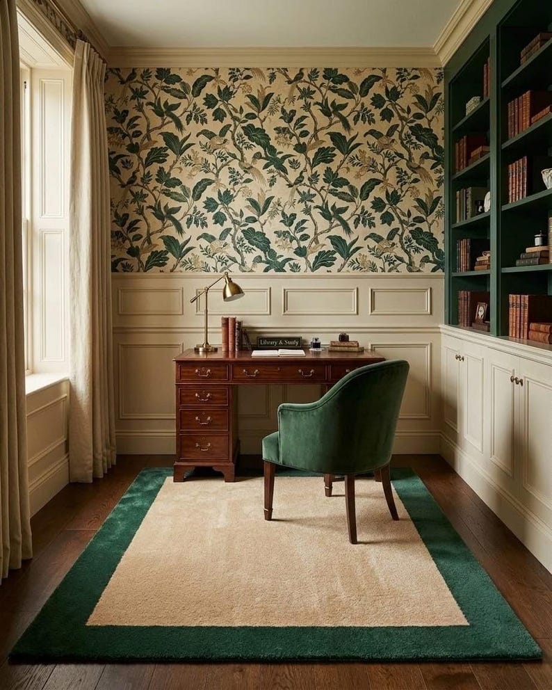 Puede incluir: Una oficina en casa con un escritorio de madera oscura con cajones, un sill&oacute;n de terciopelo verde y una alfombra rectangular con centro beige y borde verde. La habitaci&oacute;n incluye estanter&iacute;as verdes integradas, frisos color crema y papel tapiz floral.