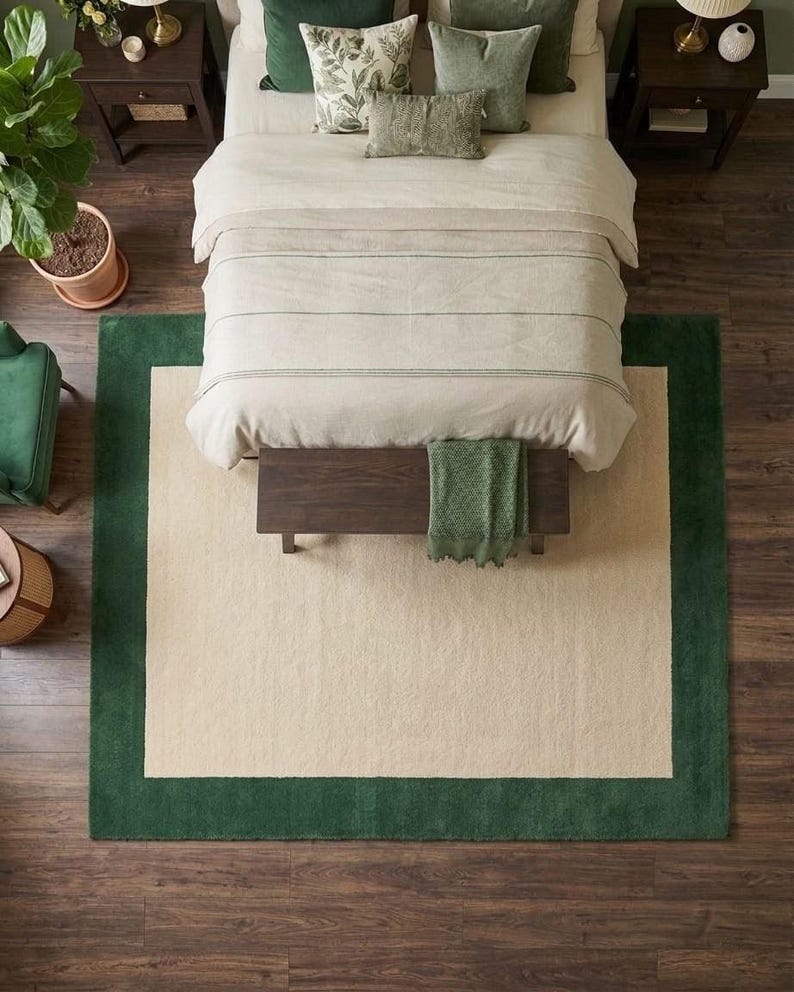 Puede incluir: Una alfombra rectangular que presenta un gran panel central de color crema enmarcado por un borde ancho de color verde oscuro s&oacute;lido. La alfombra est&aacute; colocada sobre un suelo de madera oscura bajo una cama con ropa de cama crema, un banco y cojines verdes.