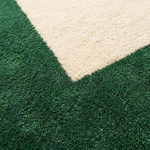 Puede incluir: Una alfombra de pelo largo con un dise&ntilde;o geom&eacute;trico que presenta un borde de color verde bosque profundo y una secci&oacute;n central en color crema. La textura es gruesa y afelpada, mostrando una clara transici&oacute;n diagonal entre los dos colores.