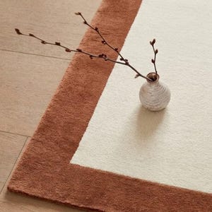 Puede incluir: Una alfombra rectangular con un gran panel central de color crema bordeado por un marco ancho y texturizado de color terracota. Un pequeño jarrón de cerámica de color blanco roto con una rama fina de color marrón oscuro descansa sobre ella.