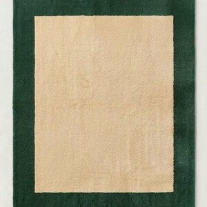 Puede incluir: Una alfombra rectangular que presenta un panel central de color beige s&oacute;lido rodeado por un borde ancho de color verde bosque oscuro. La alfombra tiene una superficie de pelo texturizado y afelpado que crea un aspecto suave.