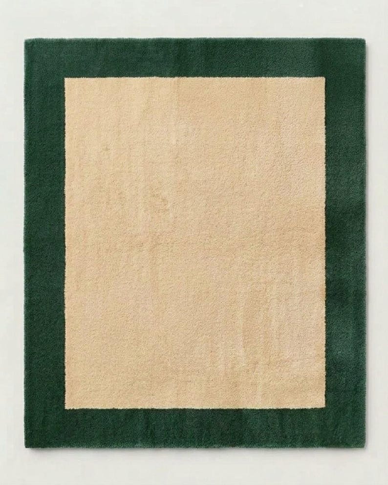 Puede incluir: Una alfombra rectangular que presenta un panel central de color beige s&oacute;lido rodeado por un borde ancho de color verde bosque oscuro. La alfombra tiene una superficie de pelo texturizado y afelpado que crea un aspecto suave.