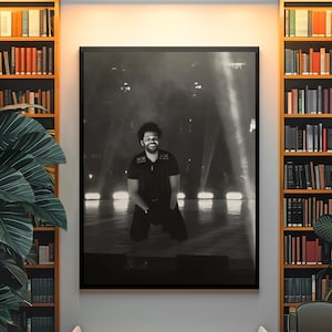 May include: A black and white framed wall art print featuring a person kneeling on a stage under bright spotlights. The person is wearing a dark short-sleeved shirt and dark pants. The image is displayed in a room with bookshelves and a bench.