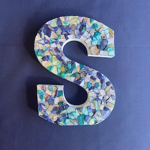 May include: A decorative letter S covered in a mosaic of small, irregular pieces of stone and shell in shades of blue, teal, turquoise, and white. The letter has a white base and a smooth, flat surface, with the colorful fragments arranged closely.
