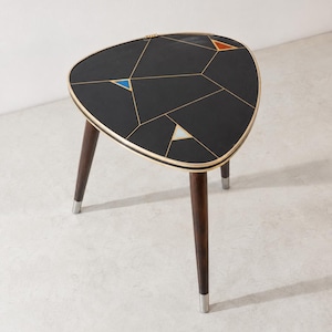 Puede incluir: Una mesa auxiliar de tres patas con un tablero triangular negro que presenta líneas geométricas doradas y pequeños triángulos incrustados en azul, azul claro y naranja. La mesa tiene patas cónicas de madera marrón oscuro con pies plateados.