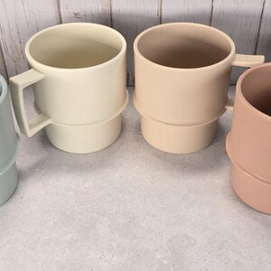 May include: Four stackable mugs in a row, each featuring a distinct matte finish. The colors from left to right are sage green, cream, light beige, and dusty rose. Each mug has a cylindrical body with a horizontal ridge and a square-shaped handle.