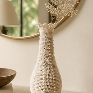 May include: A tall, cream-colored crochet vase featuring a textured, hand-stitched pattern adorned with small, evenly spaced white pearls. The vase has a narrow neck with a scalloped, ruffled rim and contains a sprig of dried white baby's breath flowers.