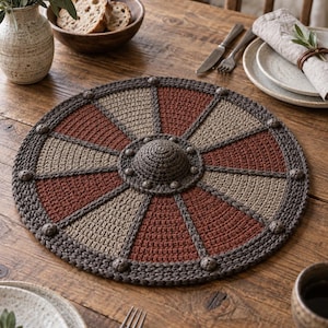 Op de afbeelding: Een ronde gehaakte placemat met een radiaal patroon van afwisselende donkerbruine, lichtbeige en terracotta rode segmenten. Het ontwerp bevat een verhoogde centrale koepel en kleine decoratieve knoppen langs de buitenste rand.