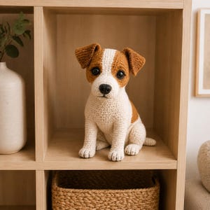 Pode incluir: Um peluche de croché em forma de cão Jack Russell Terrier. O brinquedo apresenta um corpo branco com manchas castanhas nas orelhas e na cabeça, olhos de plástico preto e um nariz cosido a preto, sentado na vertical com acabamento texturado.