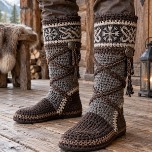 Può includere: Stivali alti all'uncinetto in marrone scuro e grigio con un risvolto decorativo caratterizzato da un motivo a intreccio bianco di ispirazione nordica e un simbolo centrale. Gli stivali presentano lacci incrociati marroni, nappe e bottoni.