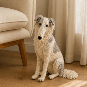 May include: A crocheted dog figurine featuring a long snout, pointed ears, and a shaggy coat in shades of cream and grey. The stuffed animal is depicted in a seated position with a textured, yarn-based construction and dark, round eyes.