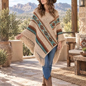 May include: A beige crochet poncho featuring a V-neckline and long fringe trim along the hem. The garment is decorated with horizontal bands of geometric patterns in shades of turquoise, brown, and cream, worn over blue denim jeans and brown boots.