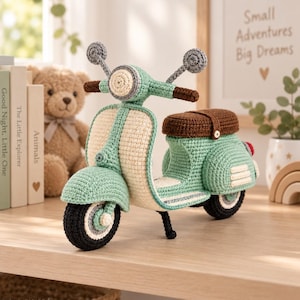 Puede incluir: Un juguete de scooter tejido a ganchillo en verde menta y crema con un asiento marrón oscuro, neumáticos negros y espejos circulares grises. El scooter cuenta con un faro delantero, un soporte y pequeñas rejillas de ventilación blancas.