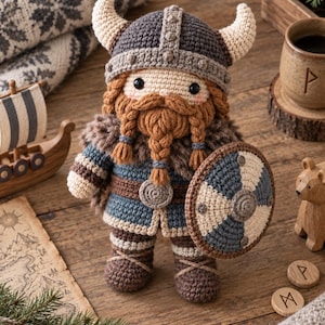 Puede incluir: Una muñeca vikinga de ganchillo con un casco con cuernos gris oscuro, una espesa barba pelirroja trenzada y una túnica azul con cinturón marrón. La muñeca sostiene un escudo redondo con segmentos azules y blancos y lleva botas marrones con cordones.
