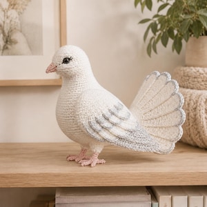 May include: A handcrafted crochet dove featuring white yarn for the body and head, with light gray accents on the wings and tail feathers. The bird has a small pink beak, pink feet, and dark eyes with stitched eyelashes, standing in a profile view.