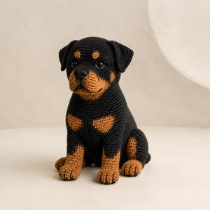 Pode incluir: Uma figura de filhote de Rottweiler feita à mão em crochê com um padrão de fio preto e castanho. O brinquedo de pelúcia tem uma superfície texturizada, olhos redondos escuros e uma postura sentada com patas detalhadas e orelhas caídas.