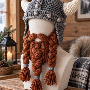 Può includere: Un elmo da vichingo all'uncinetto grigio con due corna color crema e piccoli accenti circolari, abbinato a una folta barba lavorata a maglia color arancio ruggine con tre lunghe sezioni intrecciate che terminano con nappe fissate da fasce grigie.