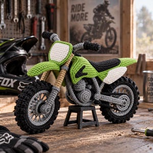 Op de afbeelding: Een gehaakte crossmotor met een groene carrosserie, zwarte zitting, witte zijpanelen en zwarte noppenbanden. De motorfiets staat op een kleine zwarte standaard en is voorzien van beige voorvorken en een wit nummerbord aan de voorzijde.