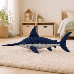 May include: A crochet swordfish toy featuring a dark blue upper body and a light gray underbelly. The plush fish has a long, pointed bill, a prominent dorsal fin, side fins, and a forked tail. It has a small black eye on the side of its head.