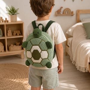 May include: A crochet turtle backpack featuring a round, sage green shell with a white and dark green hexagonal pattern. The backpack includes a small turtle head at the top, four dark green flipper-shaped side accents, and dark green straps.