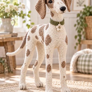 May include: A crocheted dog figurine featuring a cream-colored body with brown spots, long floppy ears, and a dark nose. The dog wears a textured olive green collar adorned with a small, white, flower-shaped charm centered with a gold bead.