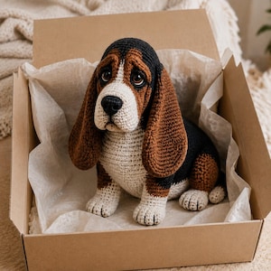 May include: A crocheted basset hound dog figurine featuring a tricolor pattern of black, brown, and cream yarn. The plush toy has long, drooping ears, a white muzzle, and dark brown eyes, sitting inside a square cardboard shipping box.