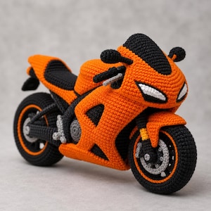May include: A handcrafted crochet motorcycle toy featuring a vibrant orange and black colour scheme. The design includes detailed black wheels with orange rims, a black seat, and white accents on the front fairing to represent the headlights.