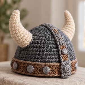 Puede incluir: Un gorro de ganchillo con forma de casco gris oscuro con dos grandes cuernos de color crema. El gorro presenta una banda marrón en la base decorada con pequeños detalles circulares grises y una franja vertical gris en la parte superior.