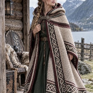 May include: A hooded crochet cloak featuring a cream-colored base with dark red and forest green geometric border patterns. The garment is secured at the neck with a circular metal brooch and worn over a solid dark green long-sleeved dress.