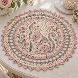 May include: A round crochet doily featuring a central cat motif in shades of cream, soft pink, and grey. The cat is surrounded by floral sprigs, enclosed within a circular border of intricate openwork stitches and a scalloped pink edge.