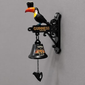 May include: A wall-mounted Guinness bell featuring a black toucan with an orange beak balancing two glasses on its head. The bracket displays the text GUINNESS ESTD 1759. The black bell is marked with GUINNESS DUBLIN and a harp-shaped pull chain.