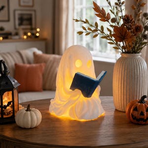 May include: A white, glowing ghost-shaped lamp featuring two circular eye cutouts and a blue book held in its hands. The lamp emits a warm yellow light from within, illuminating the ghost figure as it sits on a wooden surface alongside a lantern.