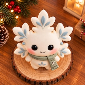 Puede incluir: Una figura con forma de copo de nieve blanco con grandes ojos negros, mejillas rosadas y detalles azules en forma de hoja. La figura lleva una bufanda de punto verde salvia con un pequeño copo de nieve blanco y un diseño de hoja en el vientre.