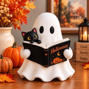May include: A white ghost figurine holding an open black book with the text Halloween, a crescent moon, and pumpkin illustrations. A small black cat with yellow eyes sits on the edge of the book. The ghost has two black oval eyes.