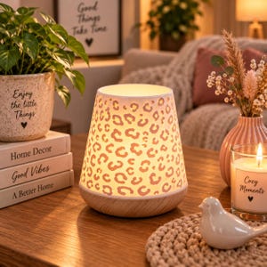 Op de afbeelding: Een conische lamp met een witte buitenkant voorzien van een uitgesneden luipaardprint en een lichte houten voet. De lamp straalt een warme gloed van binnenuit uit, waardoor het ingewikkelde dierenprintontwerp wordt verlicht.