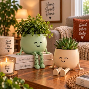 Pode incluir: Dois vasos redondos de cerâmica com rostos sorridentes e pernas articuladas pendentes. Um vaso é verde-claro com uma planta de folhas, o outro é cor de creme com uma suculenta espinhosa. Ambos estão sobre uma superfície de madeira.