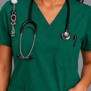 May include: A dark green V-neck scrub top featuring a chest pocket with a black stethoscope and a black pen. A decorative badge reel with a large red rectangular stone surrounded by small clear crystals is attached to the shoulder area.