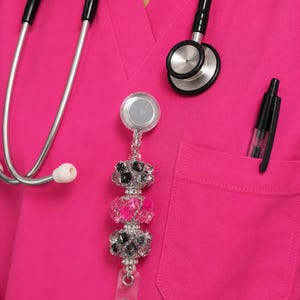 May include: A retractable badge reel featuring three decorative beads with clear rhinestones and floral accents in shades of black and pink. The reel is attached to a bright pink scrub top, which also features a black stethoscope and a black pen.
